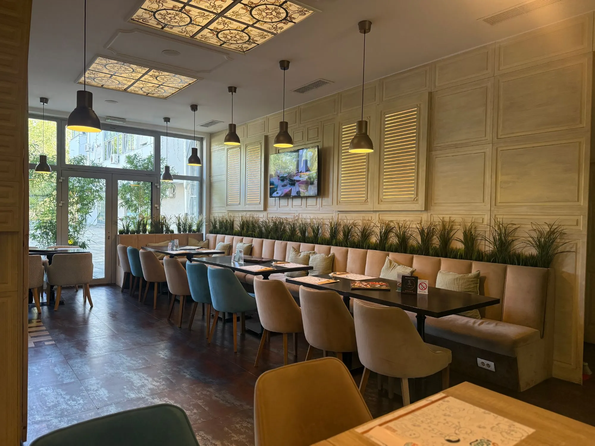 Sala porodičnog restorana Sweet & Salty na Novom Beogradu: duga tapacirana klupa sa zelenilom, viseće lampe i vitraži na plafonu